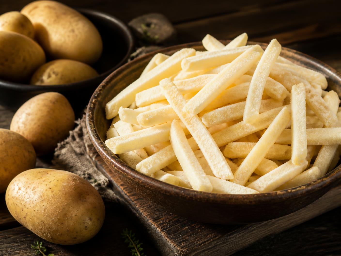 Uncooked frozen French fries in a rustic bowl beside whole potatoes
